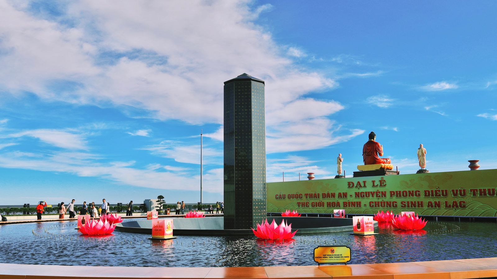 The summit terrace on the mountain: a tall dark stele in a shallow pool of pink lotus sculptures, a seated Buddha behind a painted festival banner