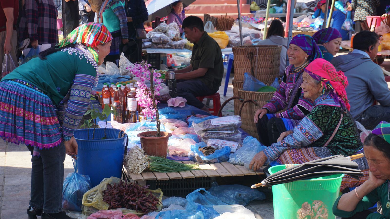 Traders in Flower Hmong headscarves and embroidered skirts sitting behind goods laid out on plastic sheeting, woven back baskets stacked beside them