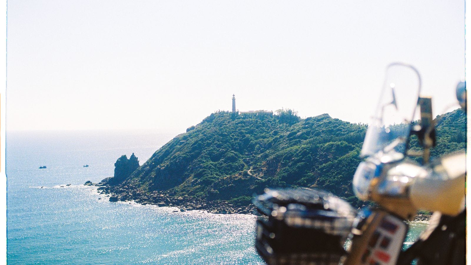 A lighthouse on a green headland above a flat blue sea, seen past the windscreen and mirrors of a parked motorbike