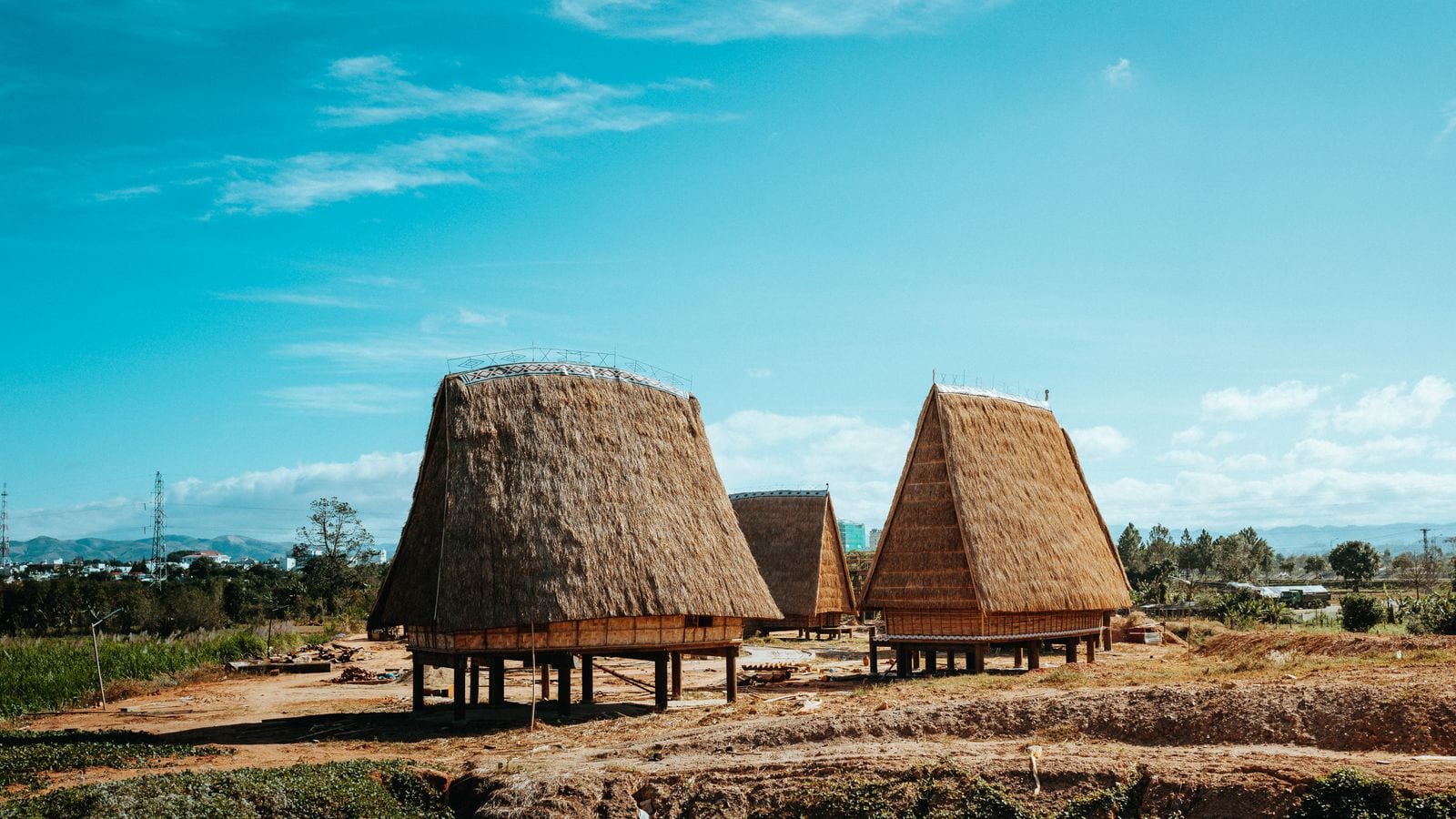 Two rong houses standing on stilts with steep thatched roofs rising to a narrow ridge, red earth around them and the town visible in the distance