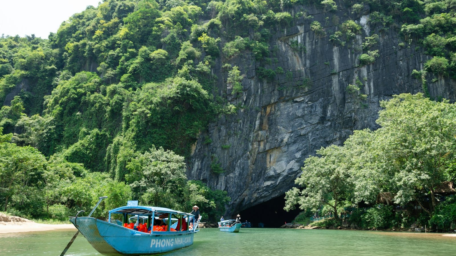 Two blue tour boats lettered PHONG NHA CAVE floating on green water at the foot of a limestone cliff, the dark cave mouth open in the rock behind them