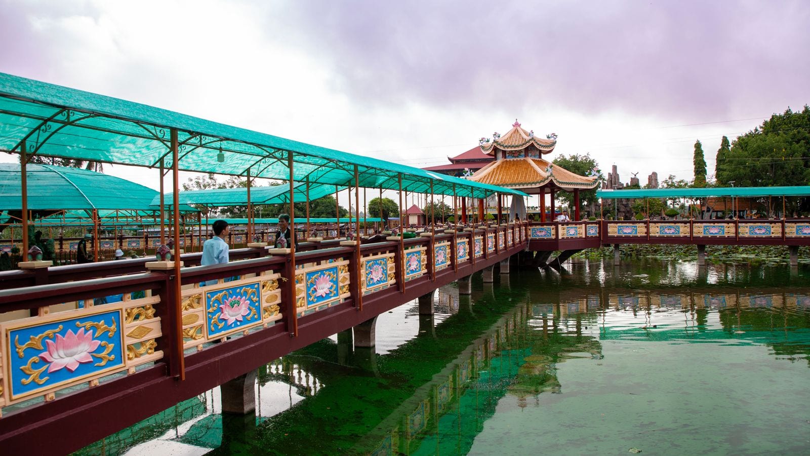 A covered walkway on stilts crossing a lotus pond, its railings panelled with painted lotus flowers, leading to a tiled pavilion