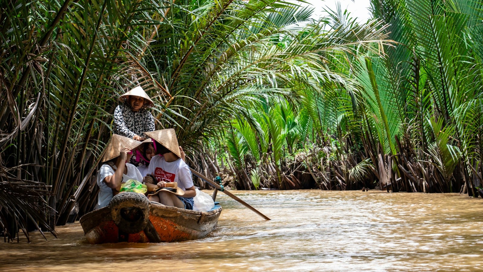 A wooden sampan being rowed up a narrow channel under nipa palms, the rower standing at the stern in a conical hat and three passengers wearing the hats handed out for the trip