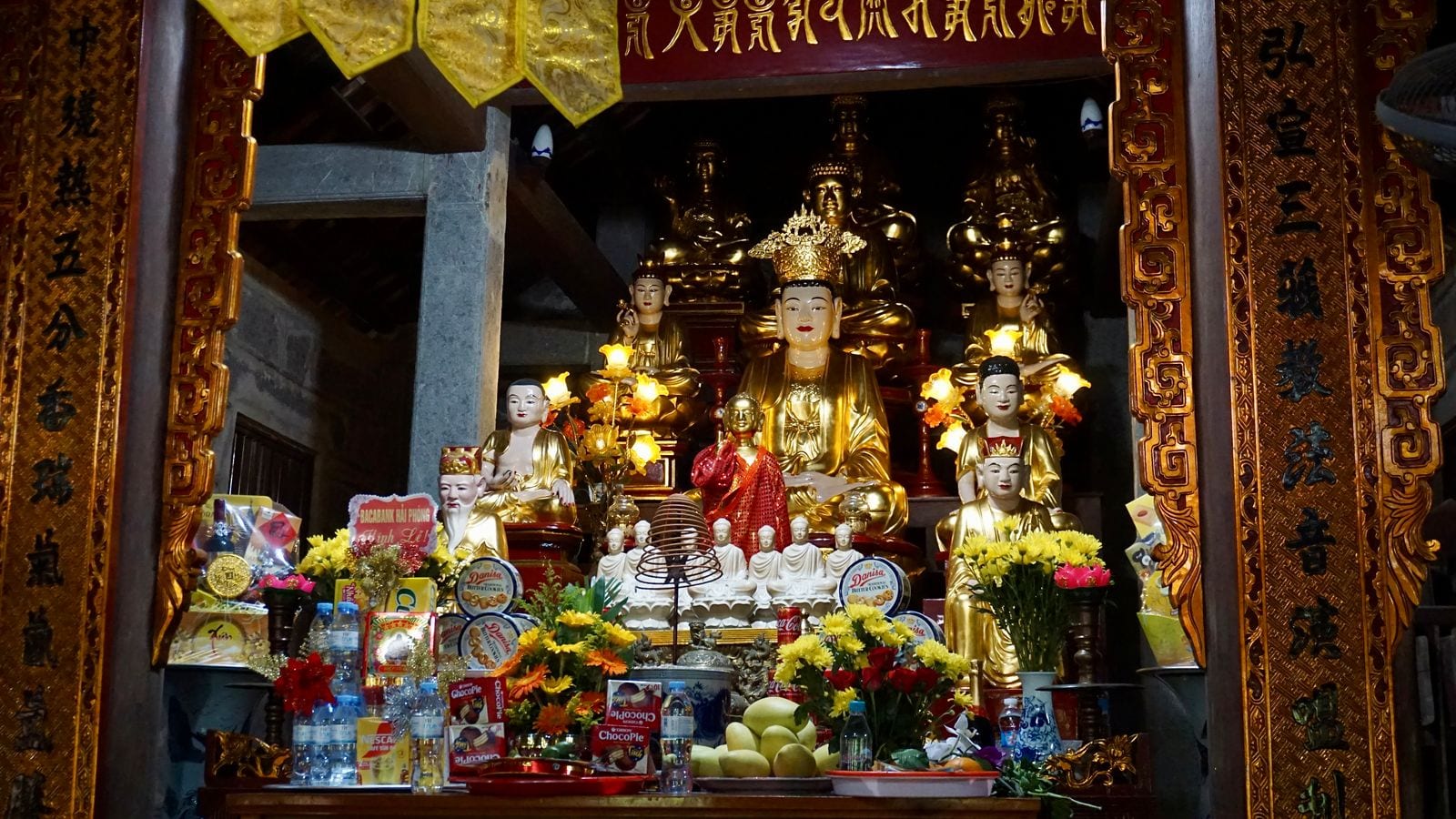 A pagoda altar of gilded and white statues banked with offerings — bottled water, biscuit tins, chrysanthemums — and a red donor banner from a Hai Phong bank propped among them