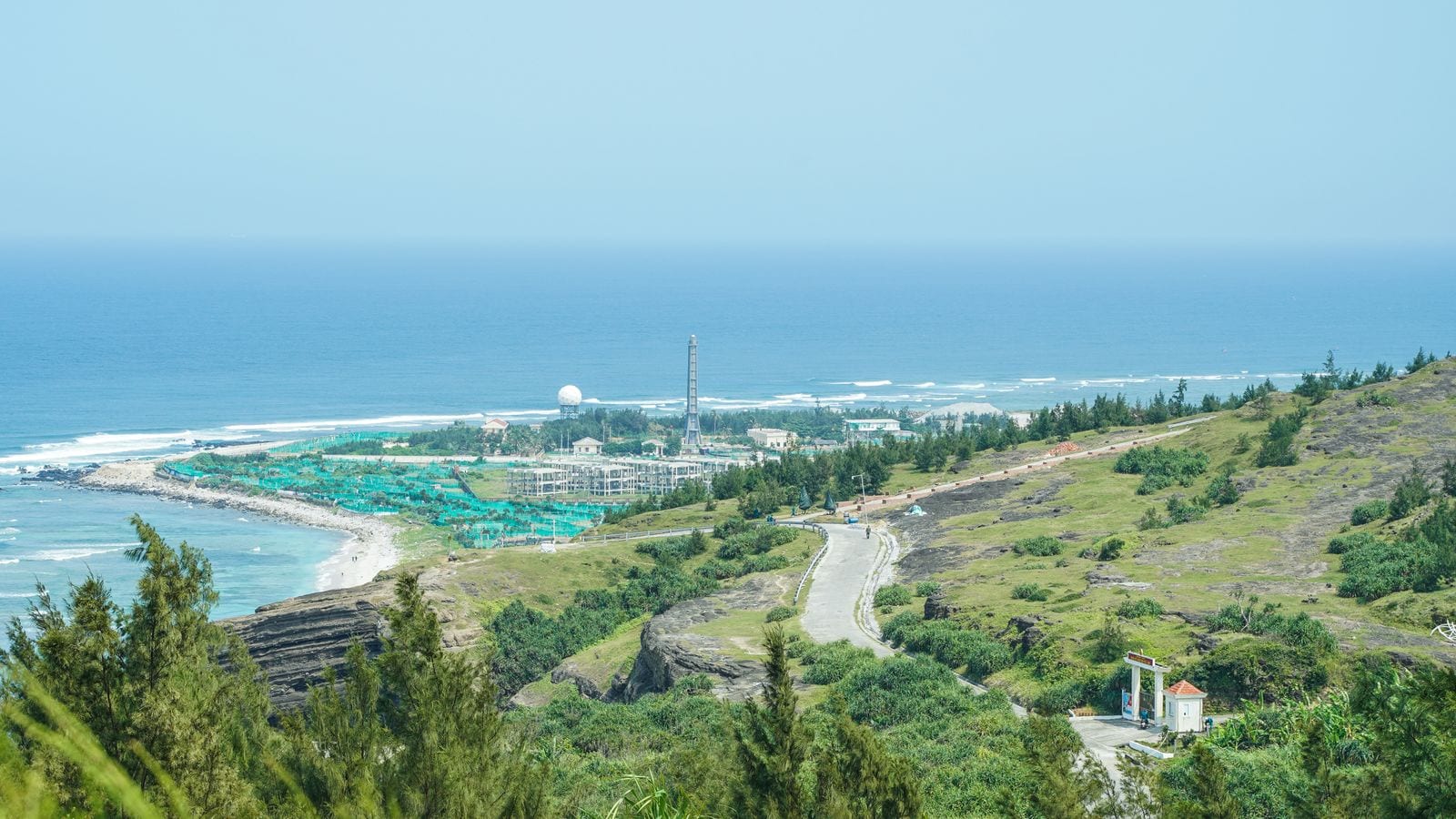 The island coast from above: a black volcanic shelf, garlic plots under green netting, a weather mast and a scatter of low buildings above a turquoise bay