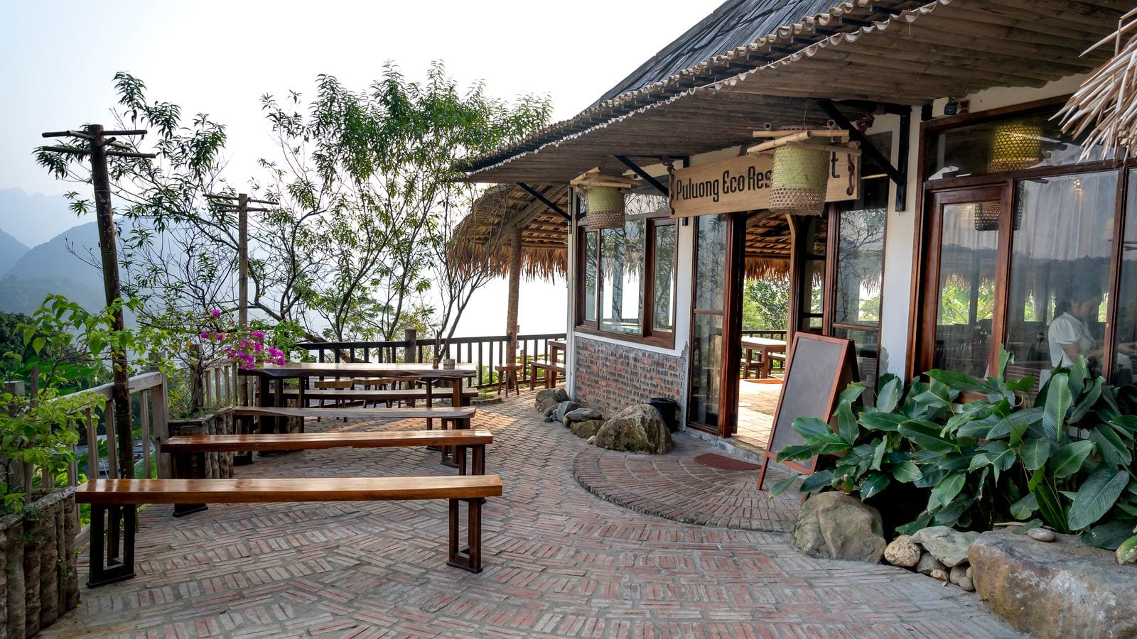 The brick terrace of an eco-lodge above the valley, long wooden benches under a thatched eave and the ridge lines fading into haze beyond the railing