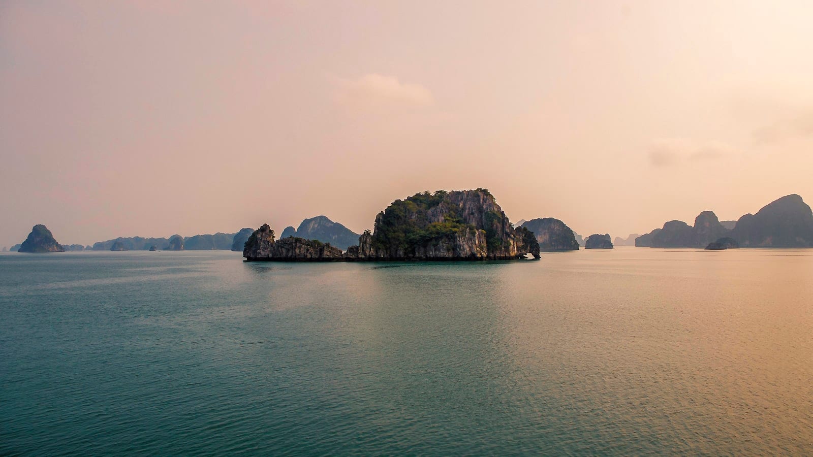 Limestone islands standing in flat pale water under a hazy sky, one large rock in the foreground
