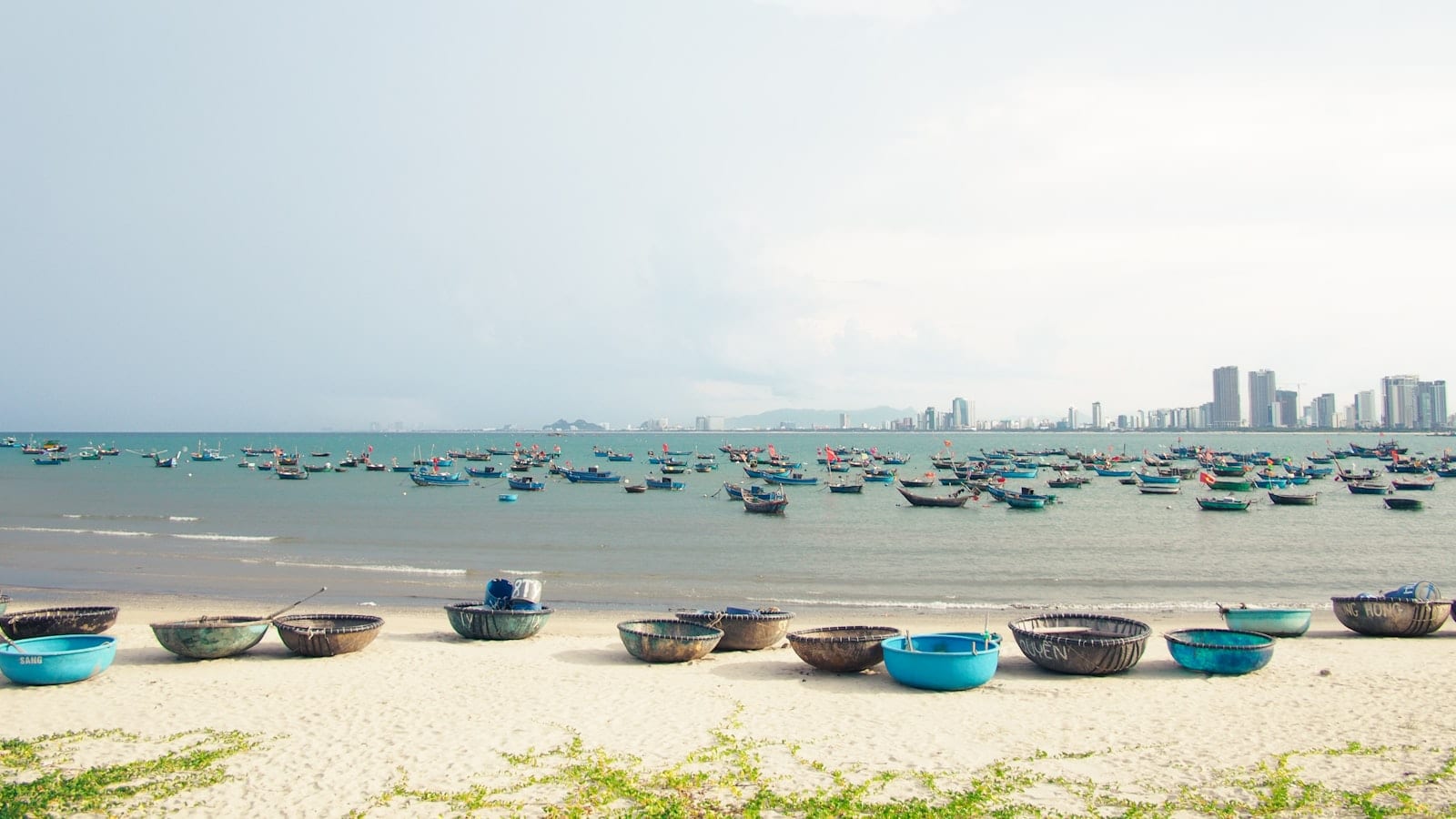 Round basket boats on white sand, fishing boats moored offshore and city towers in the distance