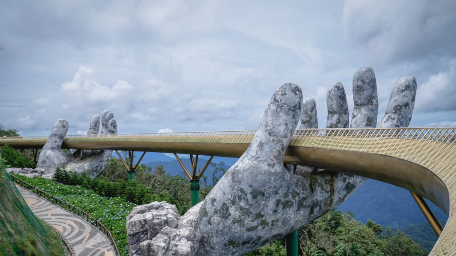 A curving gold walkway held up by two enormous weathered stone hands above forested hills