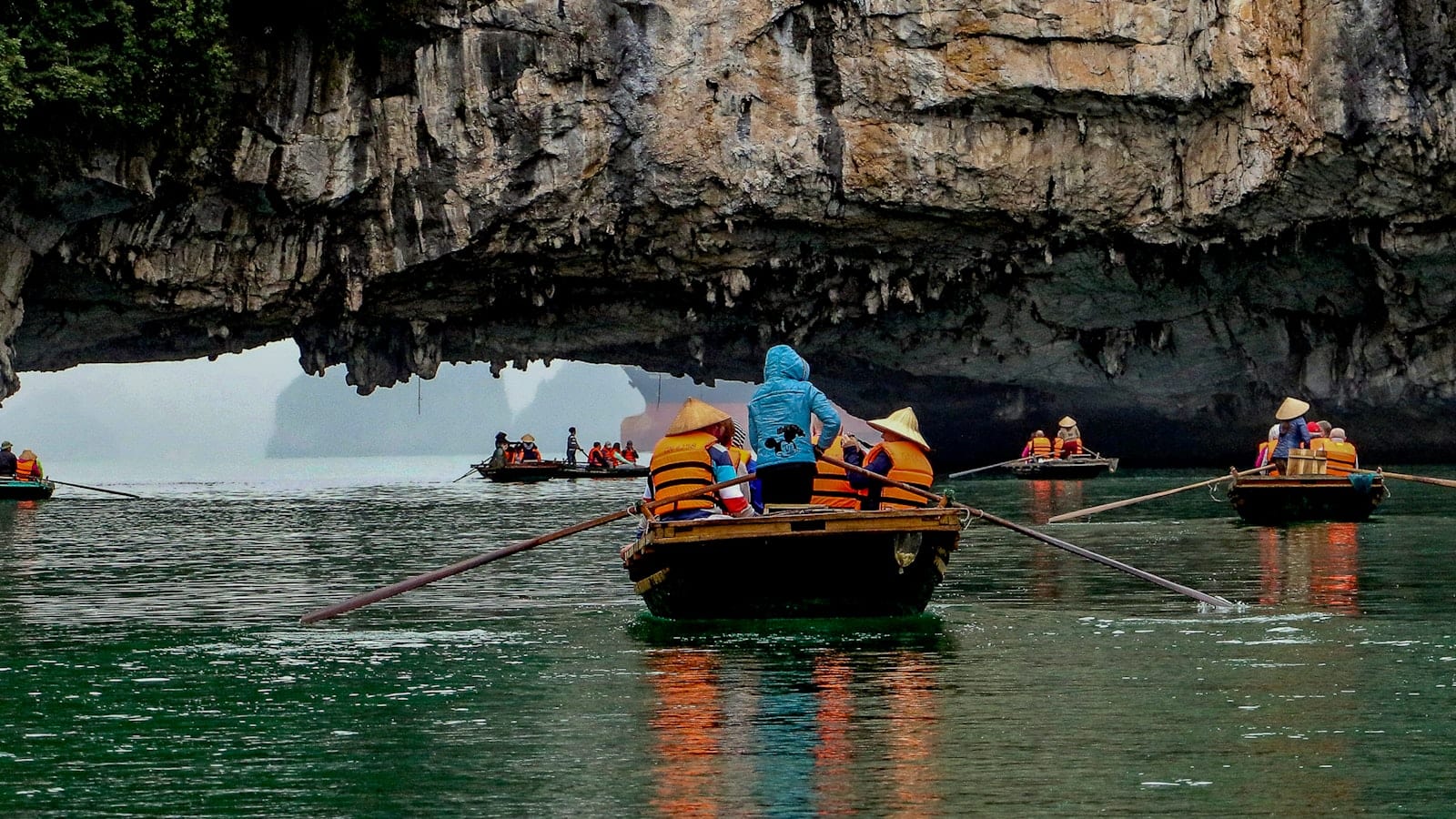 Rowing boats carrying visitors in life jackets under a low limestone arch, oars crossing the green water
