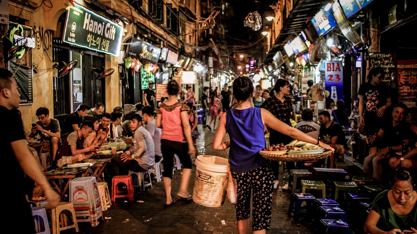 A crowded Old Quarter lane at night, plastic stools and neon signs down both sides