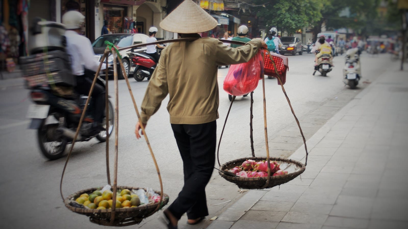 A vendor carrying fruit in two baskets on a shoulder pole along a Hanoi street