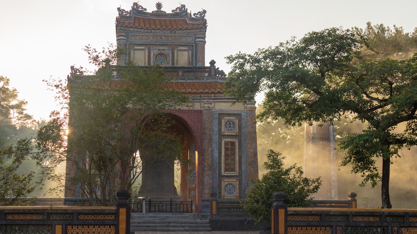 A weathered stele pavilion at a royal tomb, blue and ochre tilework under trees in morning haze