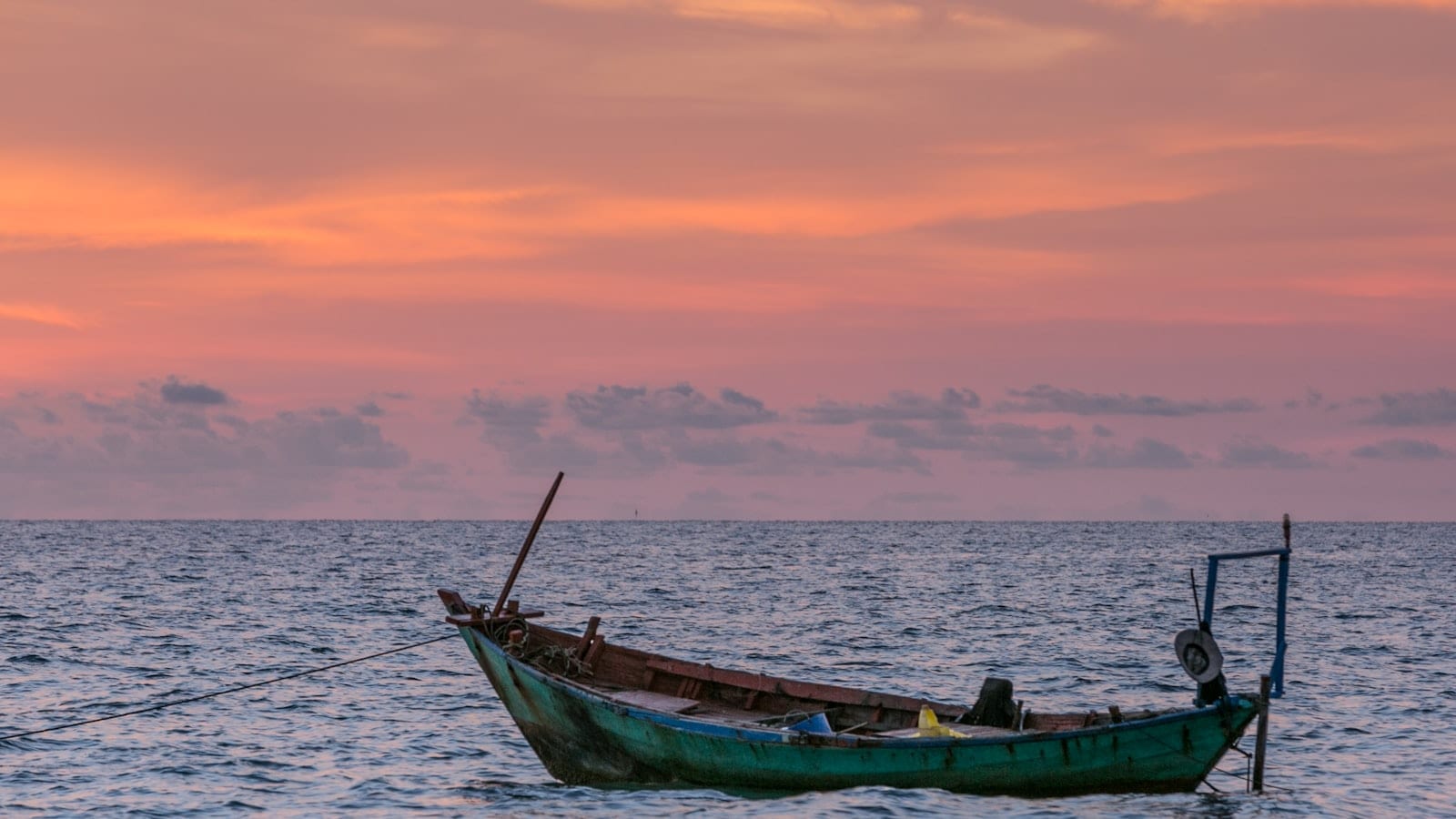 An empty green-hulled wooden fishing boat moored on flat water under a pink and orange sunset sky