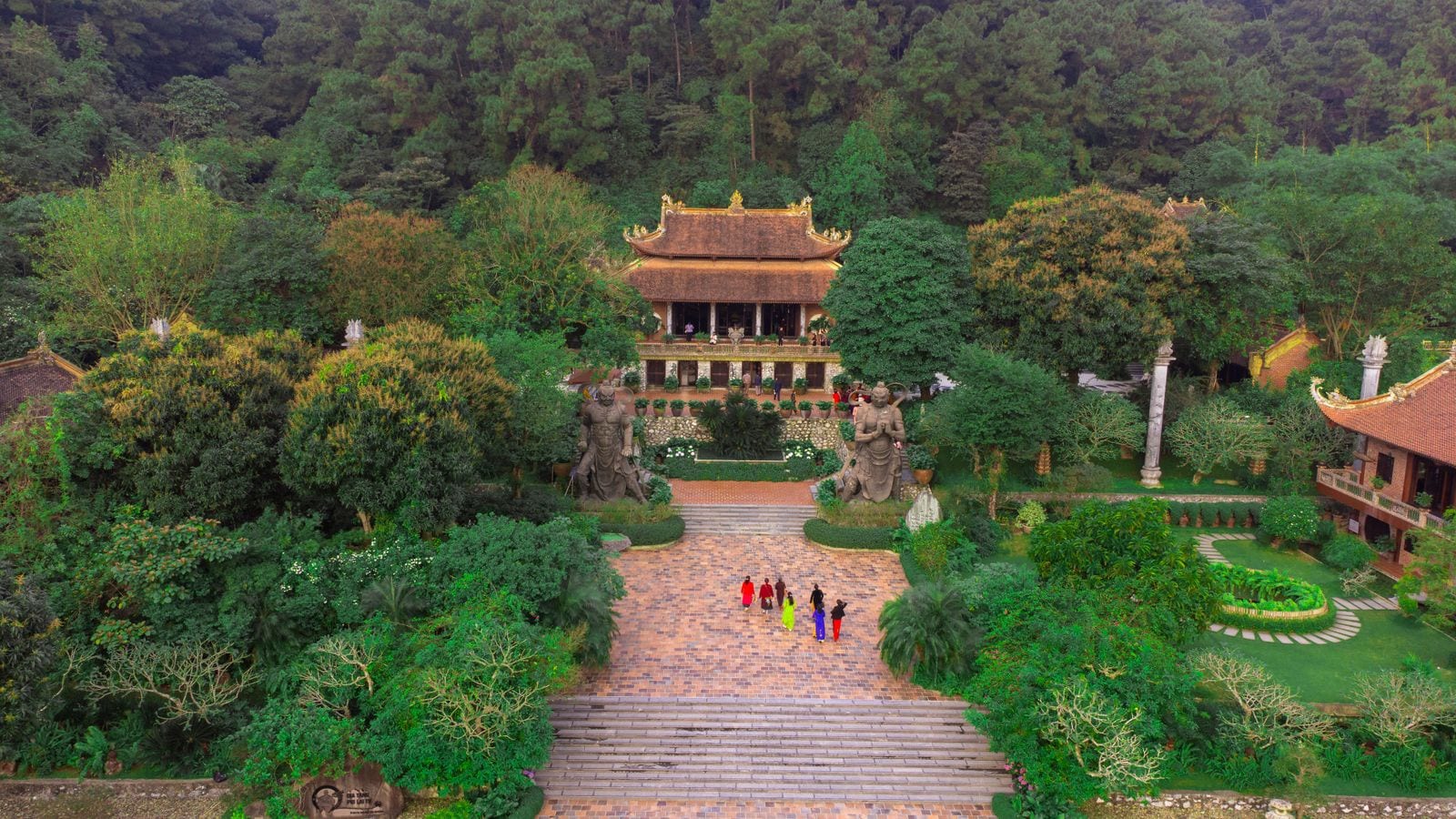 A pagoda at the head of a wide brick avenue, two carved stone guardians standing either side of the steps and forested hills rising behind the roof
