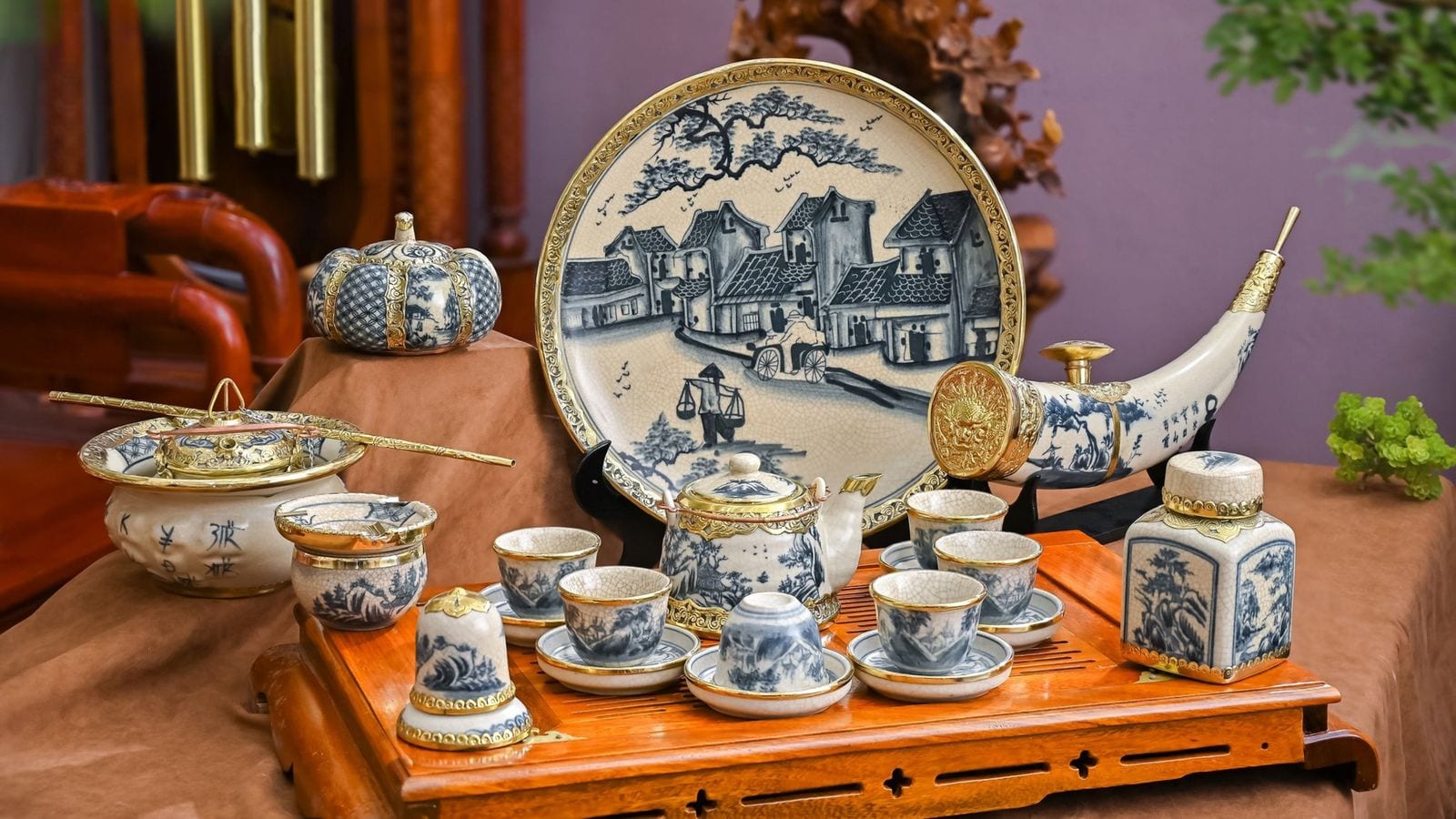 A display of blue and white pottery with gilt rims — a teapot and cups on a wooden tray, a horn-shaped vase and a large plate painted with a village street and a vendor under a shoulder pole