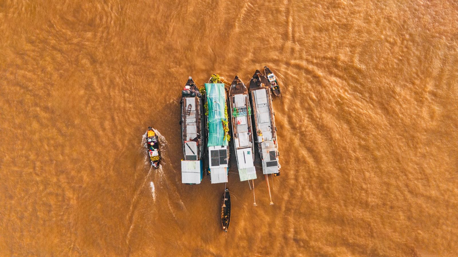 Five market boats rafted together on brown river water seen from directly above, tin cabins and solar panels on their sterns, a loaded sampan pulling away