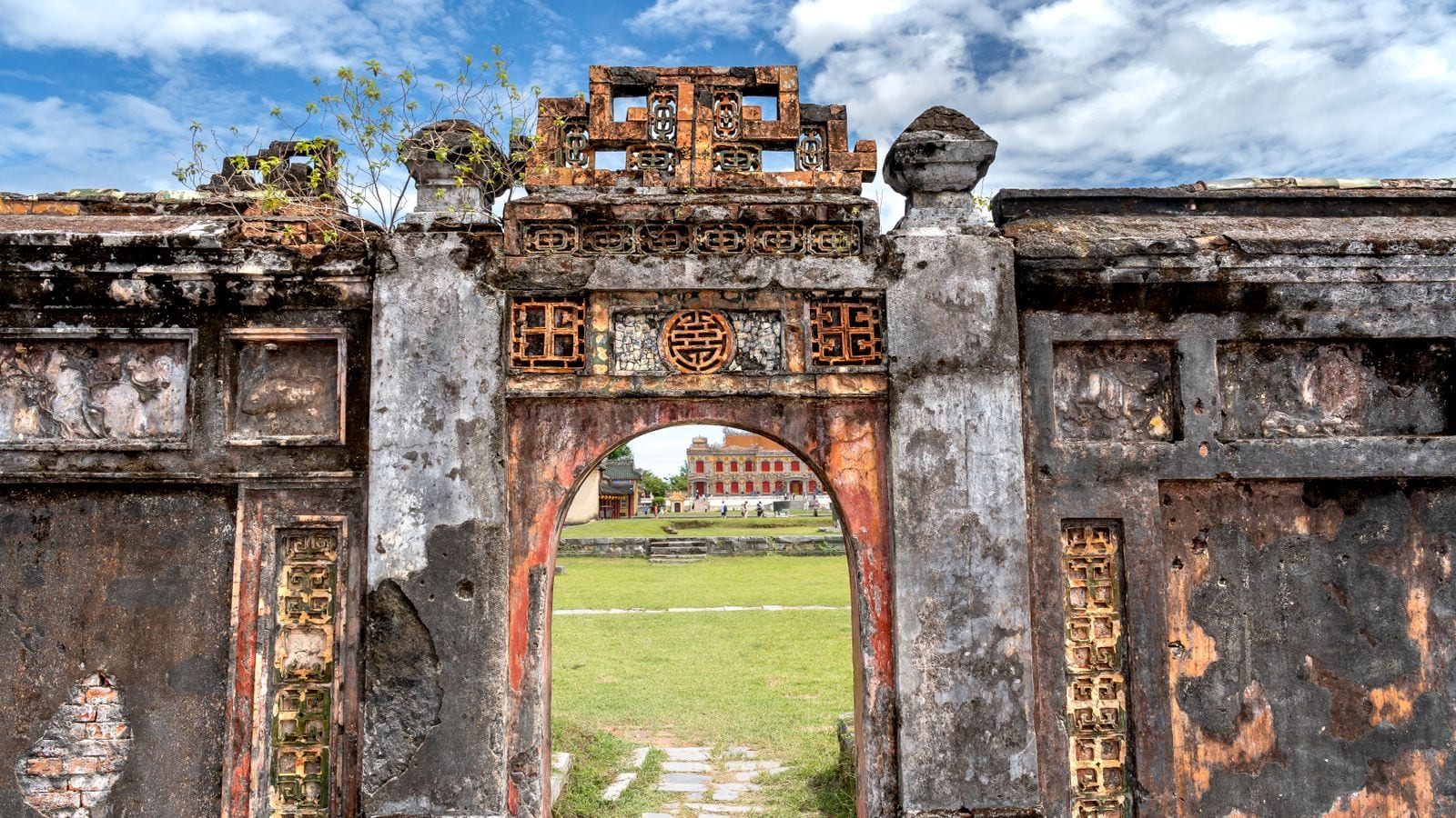 A ruined gateway in the citadel, stucco peeling off the brick and a shou roundel over the arch, with mown grass and a restored red hall visible through the opening