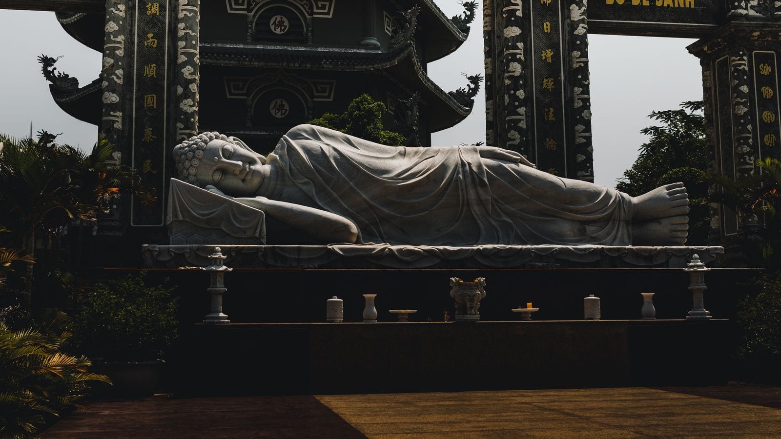 A white marble reclining Buddha on a low plinth in a pagoda courtyard, gilded columns behind it and an offering table of incense pots in front