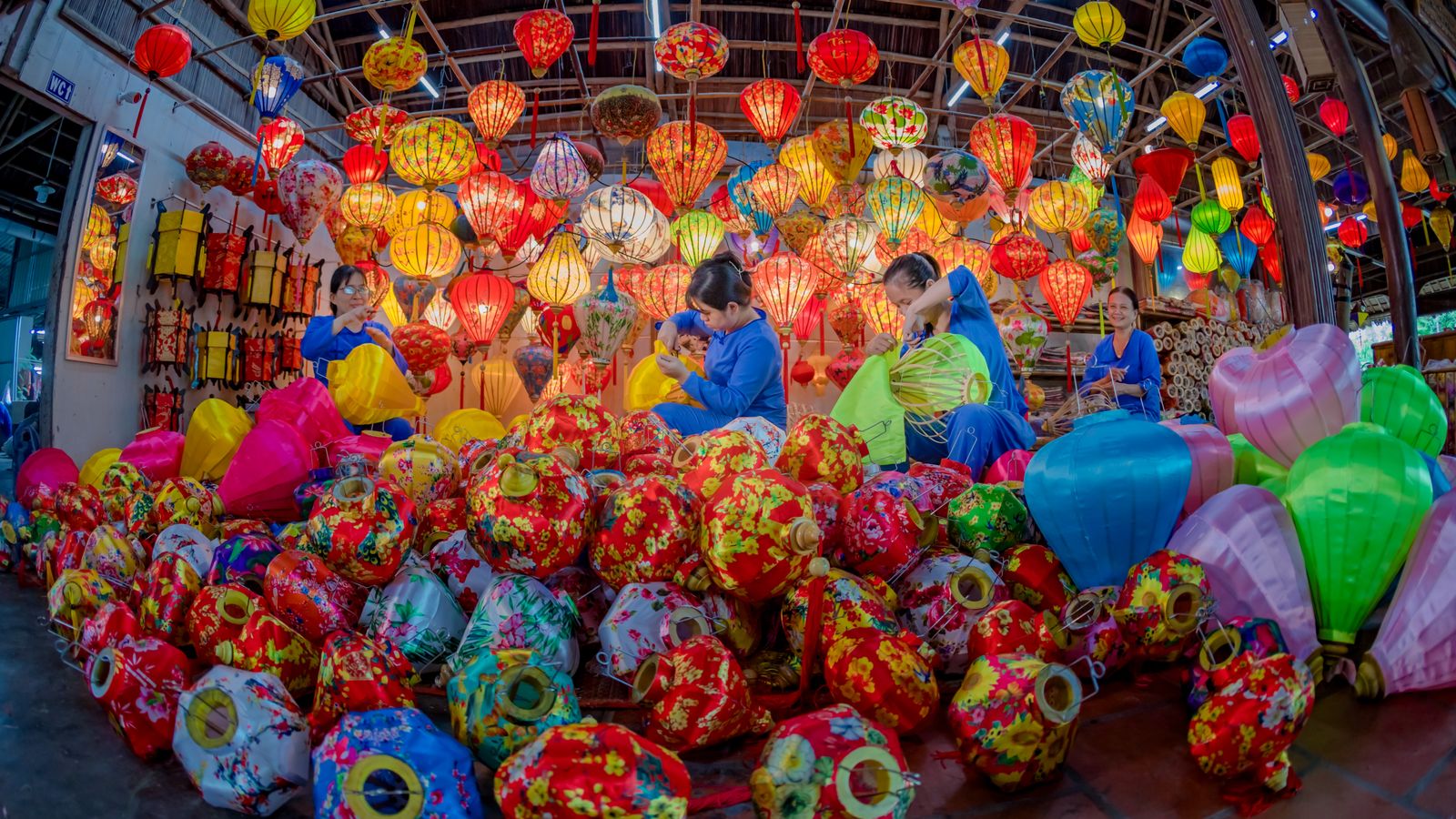 A lantern workshop with hundreds of silk lanterns hung from the roof and heaped on the floor, four women in blue shirts stretching fabric over bamboo frames in the middle of them