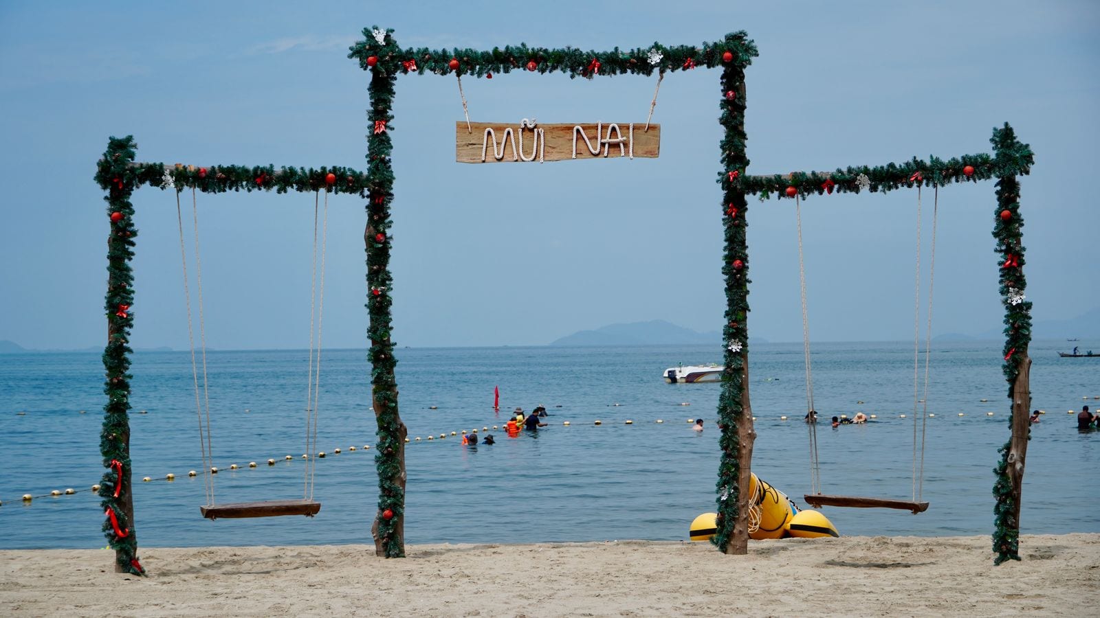 A row of beach swings hung from a decorated frame with MUI NAI lettered on a board, swimmers behind the buoy line and a hill on the horizon