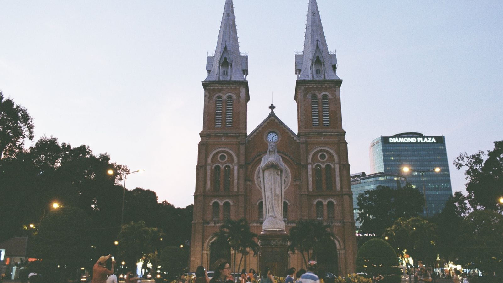The brick twin spires of the Saigon cathedral at dusk, a white statue of the Virgin on the plinth in front and the Diamond Plaza tower lit up beside it