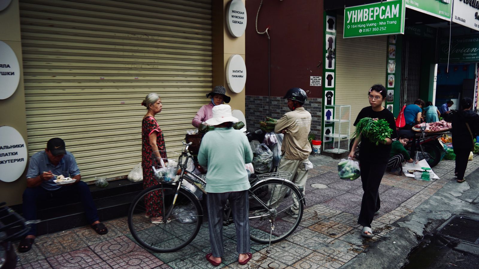 A street of shops signed in Russian — UNIVERSAM, dressing gowns, pyjamas — with a vegetable seller working the pavement in front of them and a meat stall set up on a motorbike