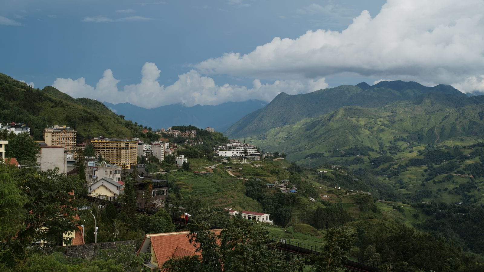 Hotel blocks stacked down a ridge above a green valley, terraces and scattered farms running away up the far side under building cloud