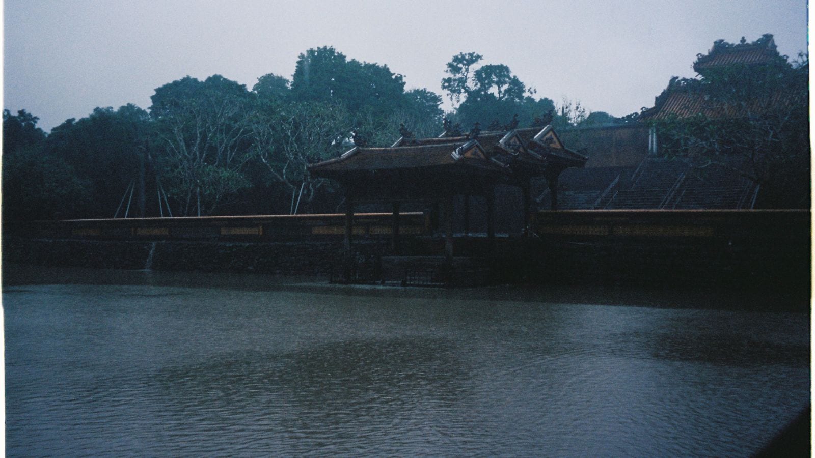 A timber pavilion with a tiled roof standing out over a tomb lake in the rain, the wall of the enclosure running away behind it