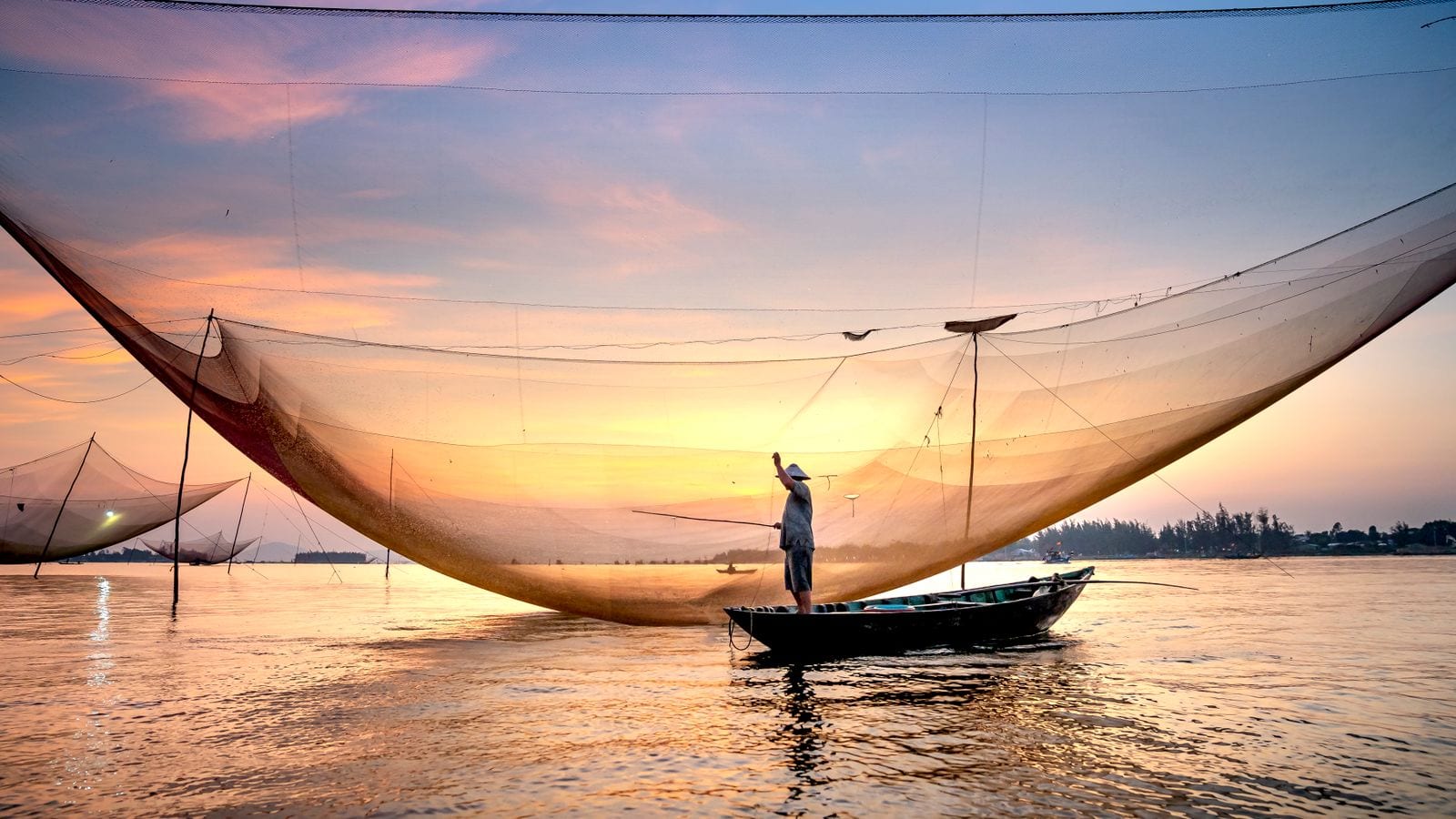 A fisherman standing in a wooden boat at dawn under the sag of a huge square lift net, more nets on their poles along the water behind him