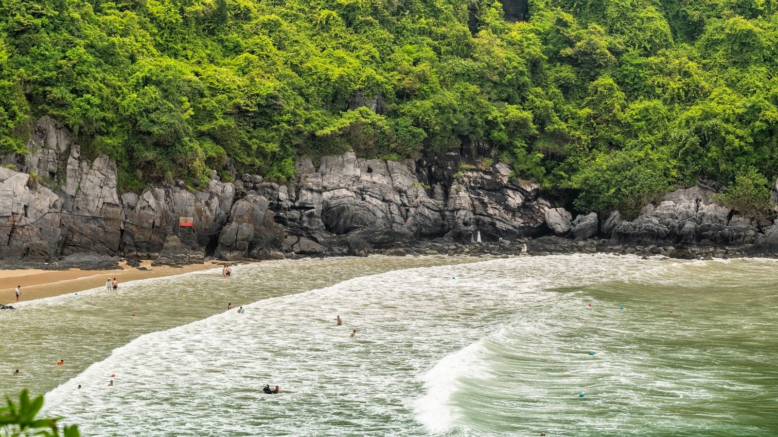 A short arc of sand under a jungle-covered limestone cliff, swimmers inside a line of buoys and a wave breaking across the mouth of the cove