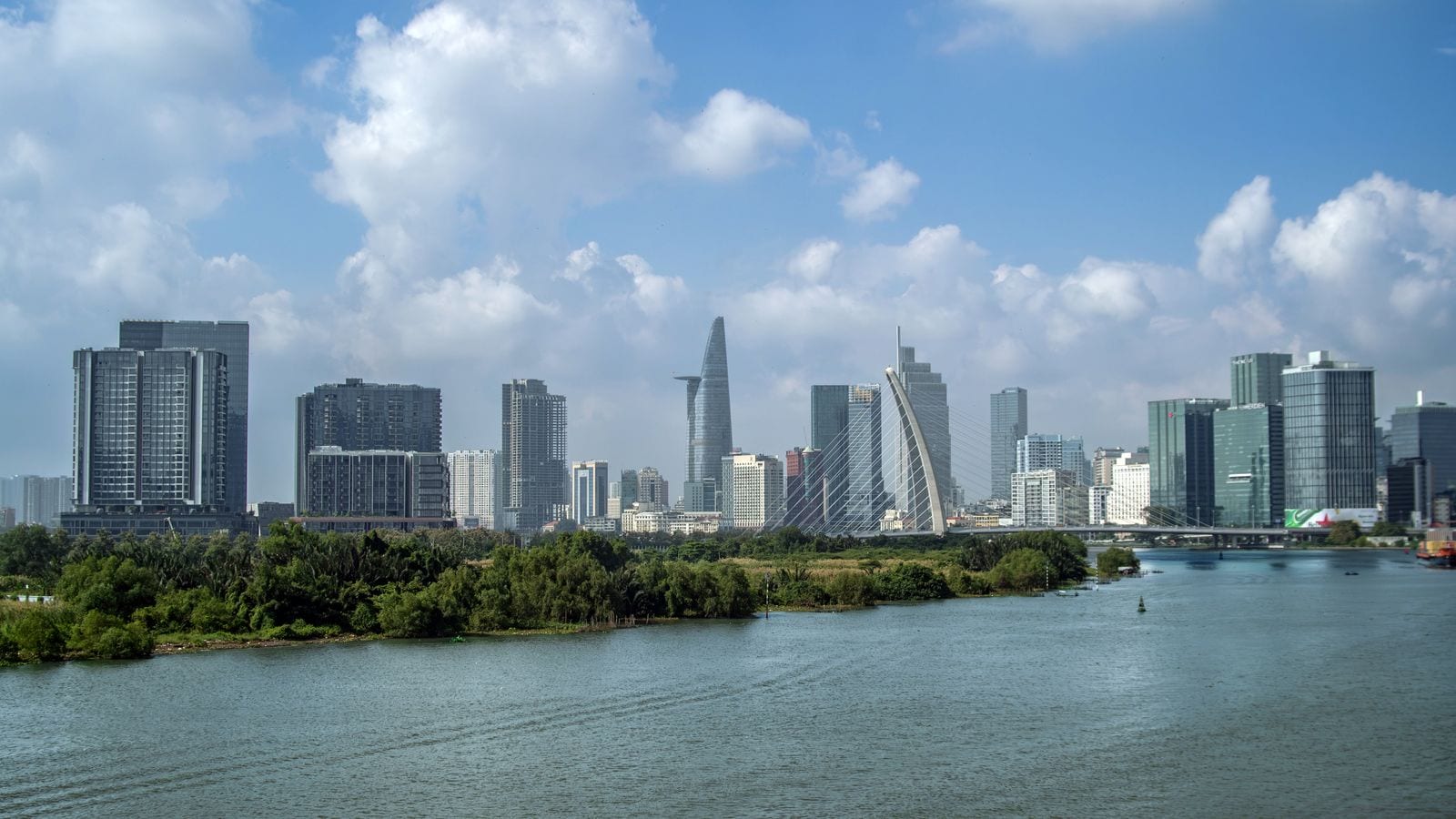 The city centre seen across the river: the tapered Bitexco tower among a wall of new blocks, the single mast of the Thu Thiem bridge to the right and scrub along the near bank