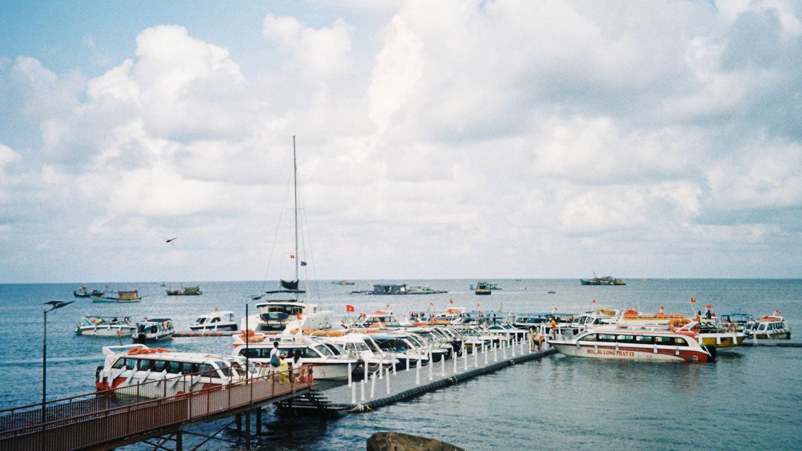 A floating pontoon lined with speedboats flying small red flags, a catamaran moored at the end of it and fishing boats out on the flat water beyond