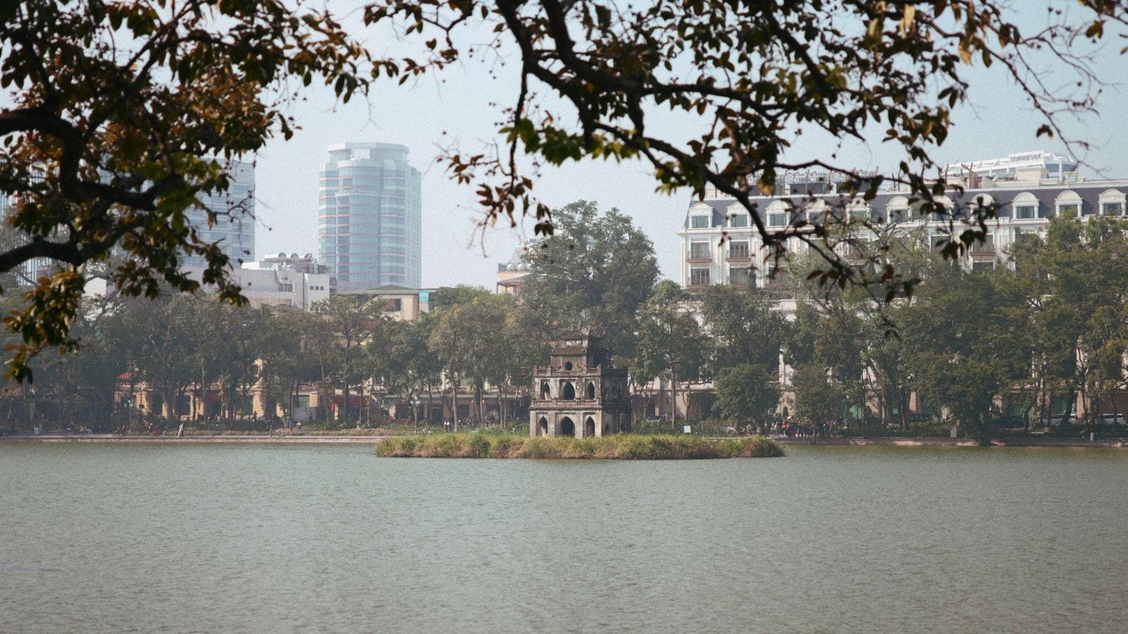 The squat stone Turtle Tower on its islet in the middle of the lake, bare winter branches across the top of the frame and shophouses and a glass office block along the far shore