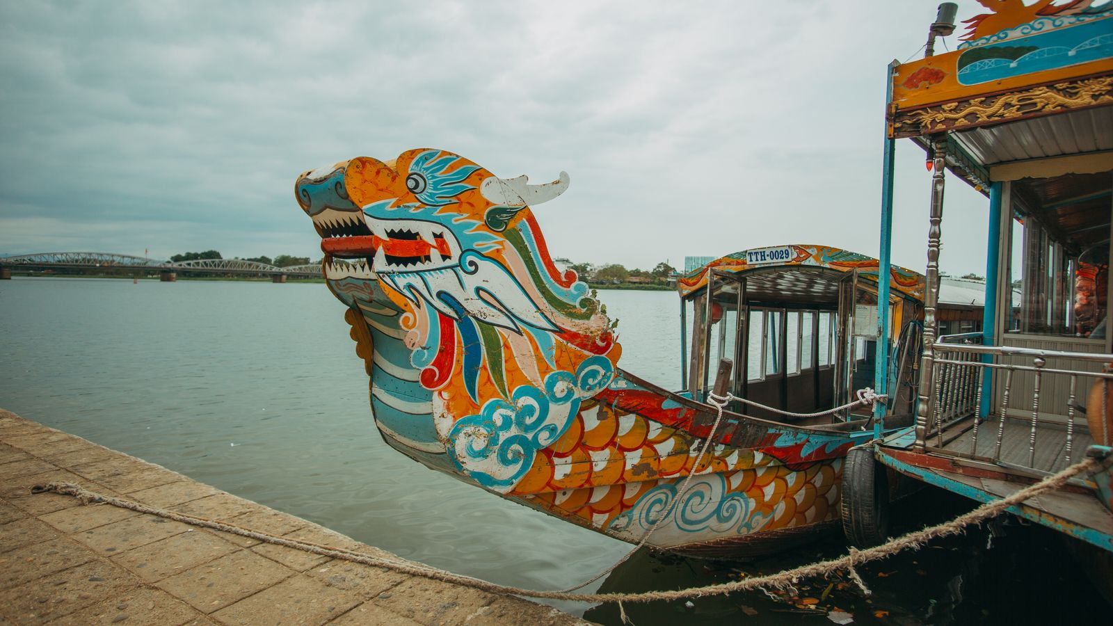 A dragon boat tied up at a stone embankment, its painted head and scaled hull filling the foreground, with the steel bridge across the river behind