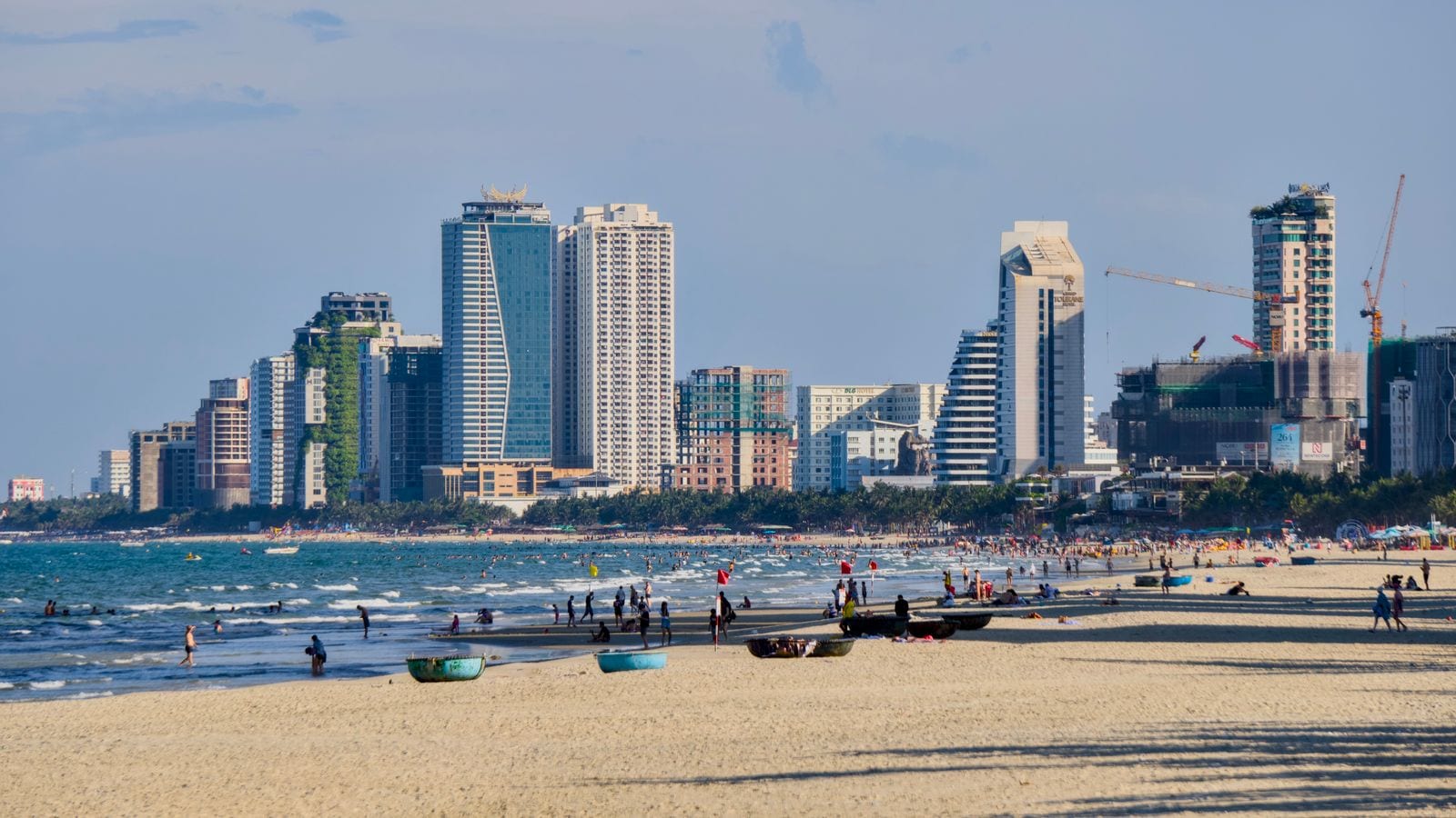 A long sand beach with a wall of hotel towers and cranes behind it, bathers in the shallows and three basket boats pulled up on the sand