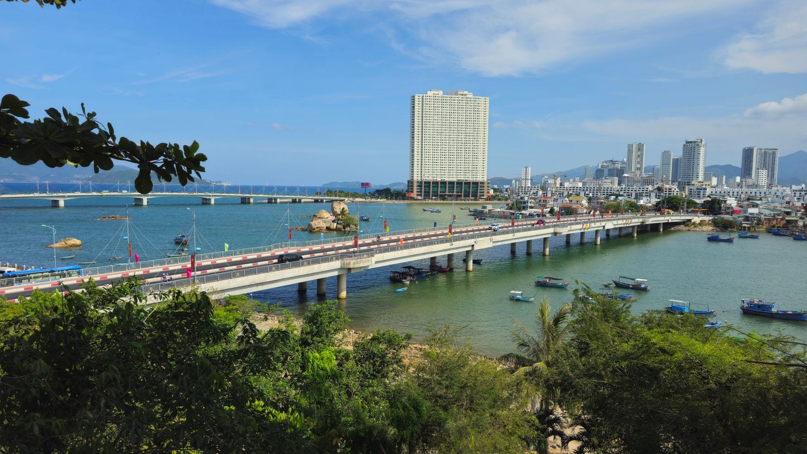 A road bridge crossing a river mouth into the city, fishing boats moored below it, a single tall hotel block on the far bank and the beach strip running away behind