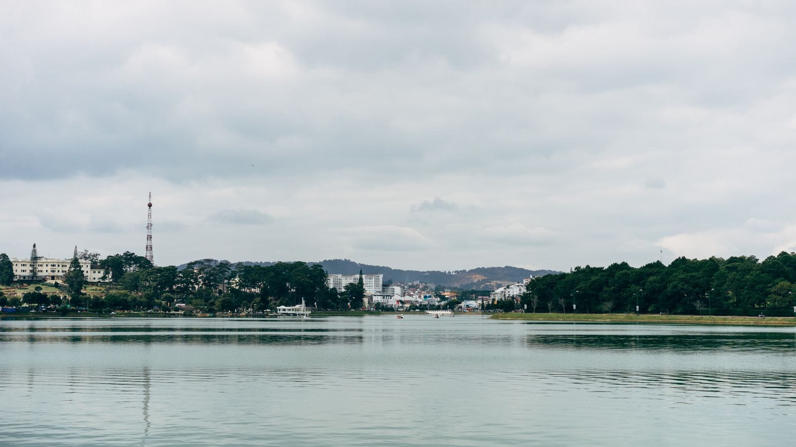 A wide lake in the middle of a hill town, pines and a radio mast on the far shore and low white buildings stepping up the slope behind them