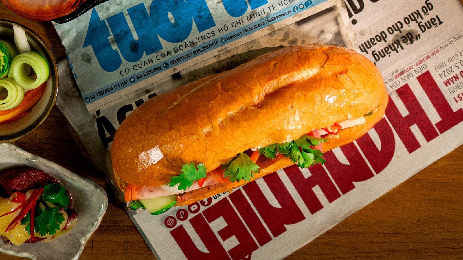 A banh mi split along one side, with pork, coriander, chilli and pickled carrot showing, resting on a folded Vietnamese newspaper