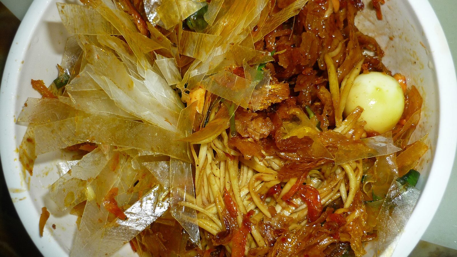 A bowl of banh trang tron from above: torn translucent rice paper, shredded green mango, dried beef in chilli oil and a peeled quail egg