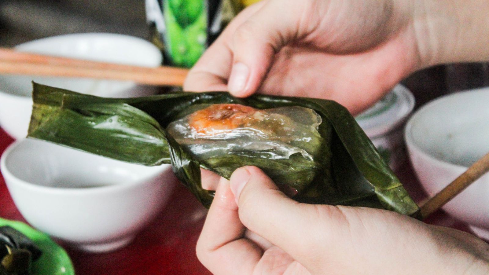 Two hands peeling back a banana leaf to show a flat, translucent banh bot loc with an orange prawn inside, small dipping bowls on a red table behind