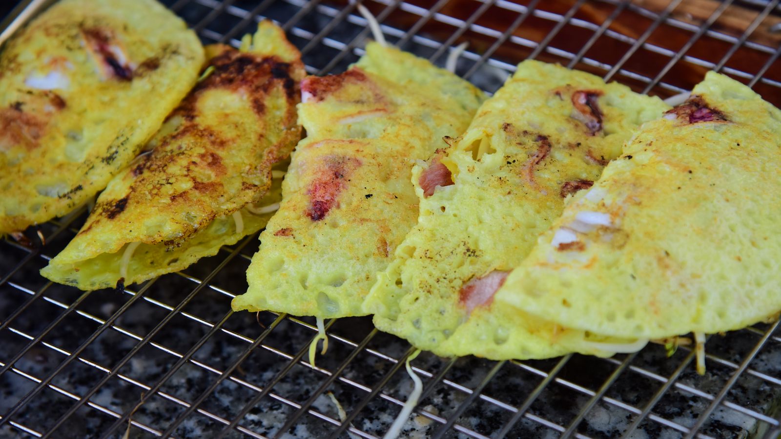 Five banh xeo folded over on a wire rack, turmeric-yellow and blistered, with prawn and bean sprouts showing at the fold