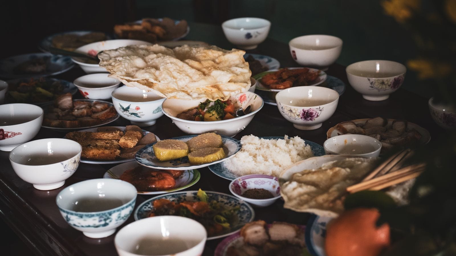 A crowded family table: a mound of plain white rice in the middle, with prawn crackers, braised pork, fried fish, giò and a small bowl at every place