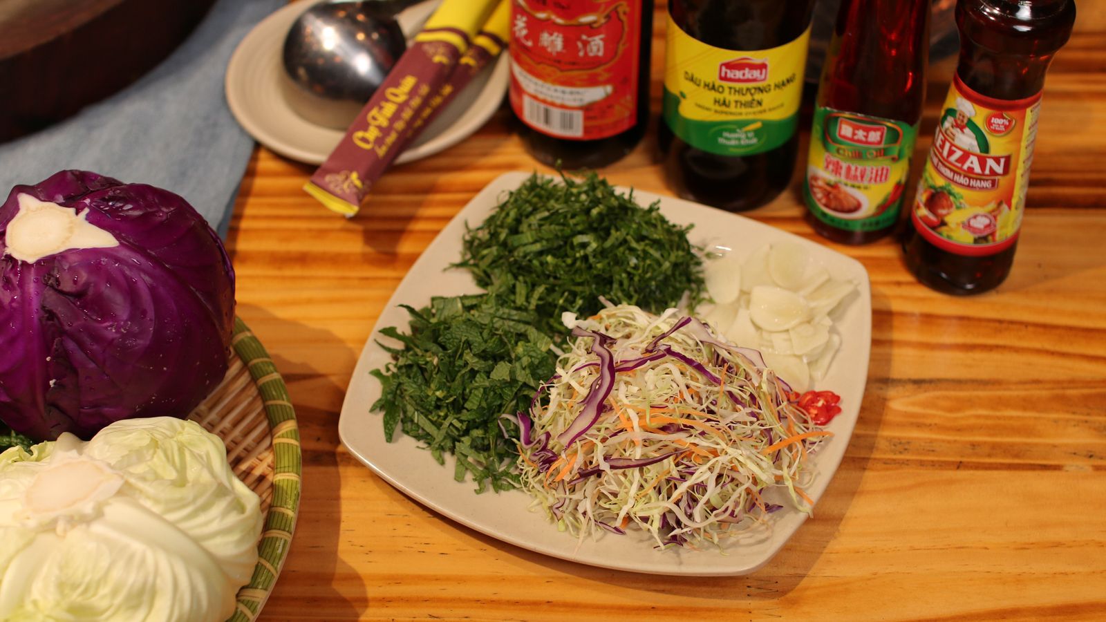 Shredded white and red cabbage with carrot on a plate, piles of chopped rau ram and mint beside it, and bottles of oyster sauce and chilli oil behind