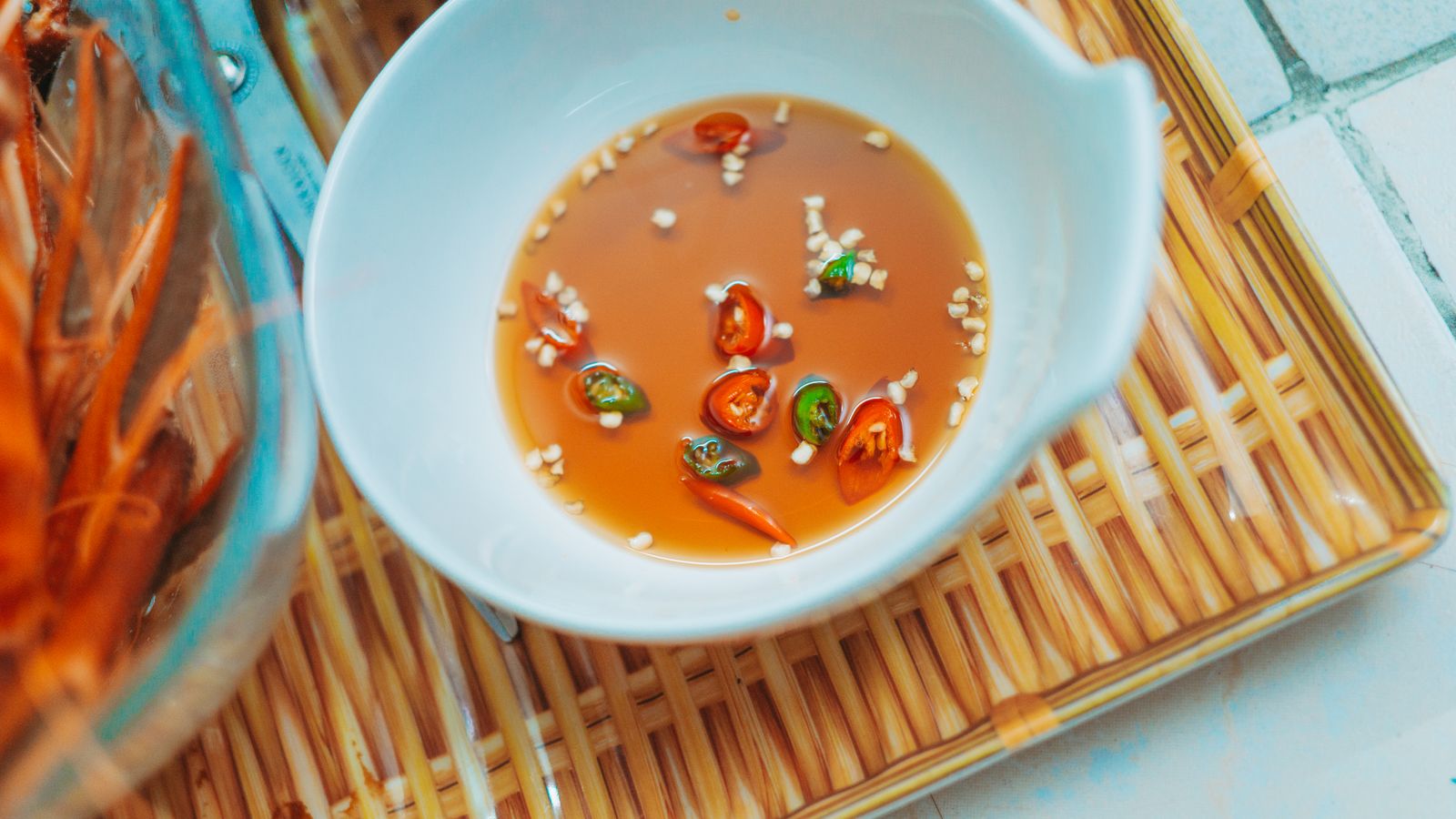 A shallow bowl of pale orange nuoc cham with sliced red and green chilli and chopped garlic floating in it, on a bamboo mat