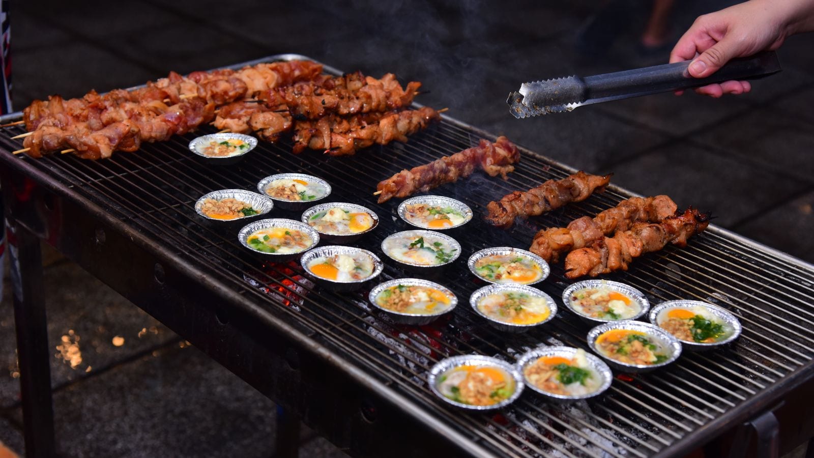 A long street grill loaded with pork skewers along the back and two dozen foil cups of egg set out over the coals in front of them, a hand reaching in with tongs