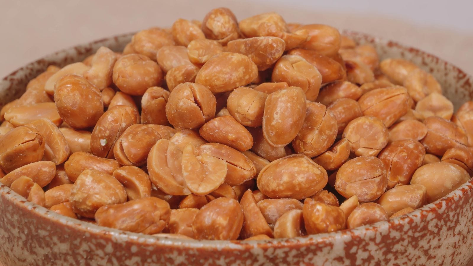 A speckled ceramic bowl heaped with roasted peanuts, some split in half, filling the whole frame