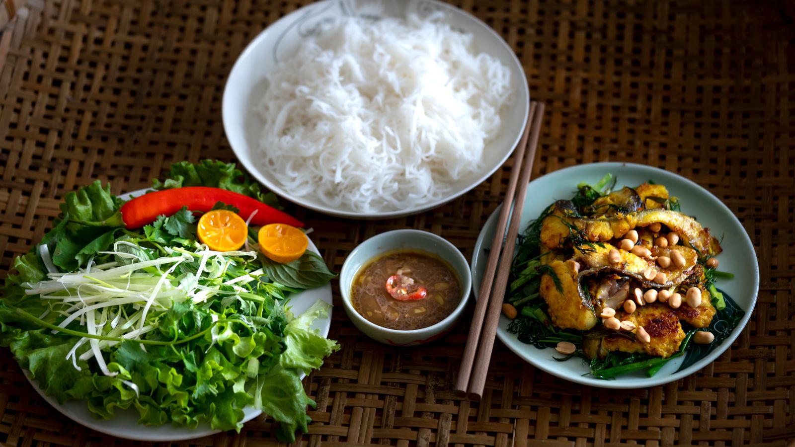 A plate of turmeric-yellow fish with dill, spring onion and peanuts, set out with a bowl of bun, a plate of herbs and a saucer of dipping sauce