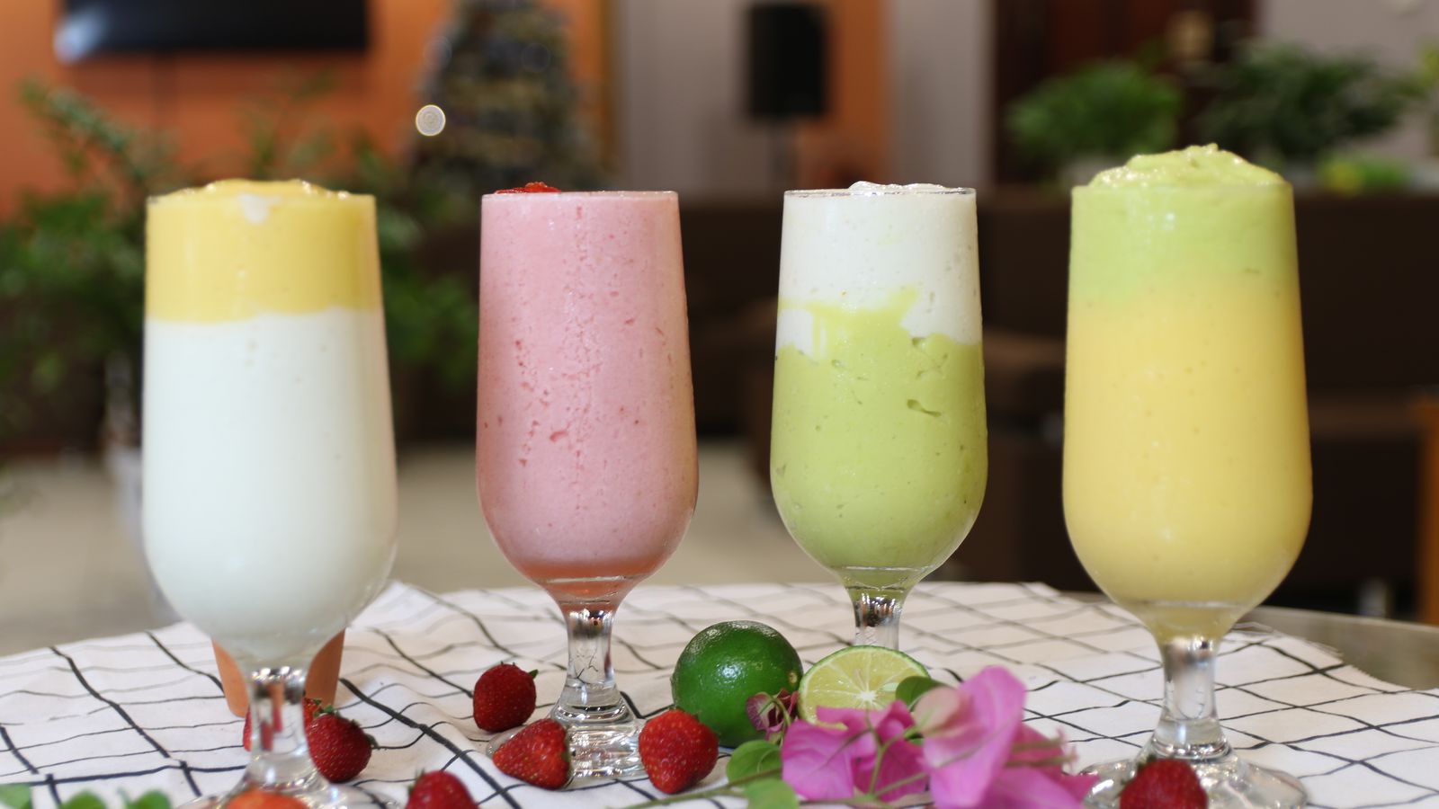 Four sinh to in tall glasses — avocado, mango, strawberry and a green one — with limes and strawberries on the cloth in front