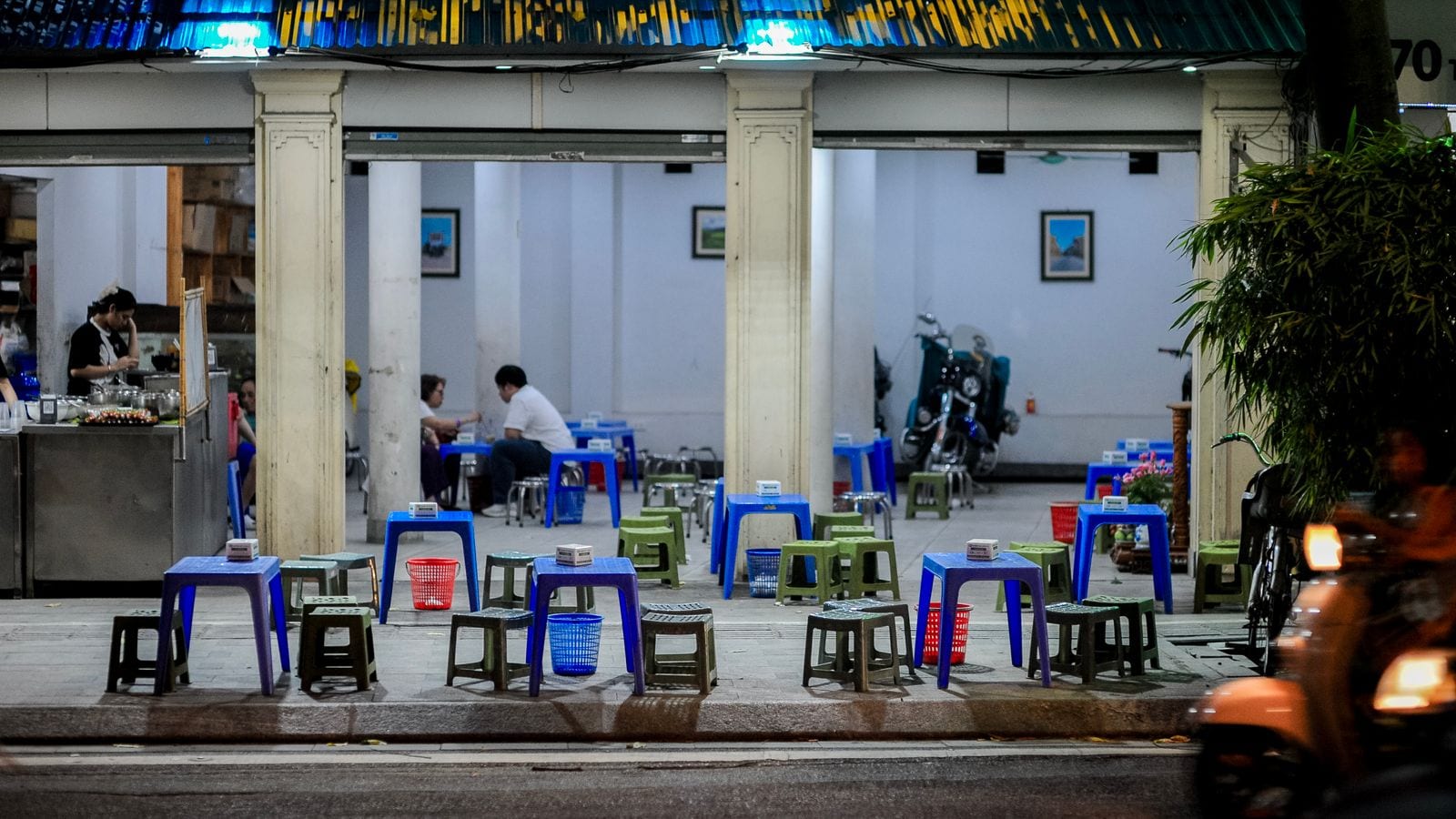 A pavement eatery at night: low blue and green plastic tables and stools set out across the flagstones under an arcade, a few diners inside and a motorbike passing